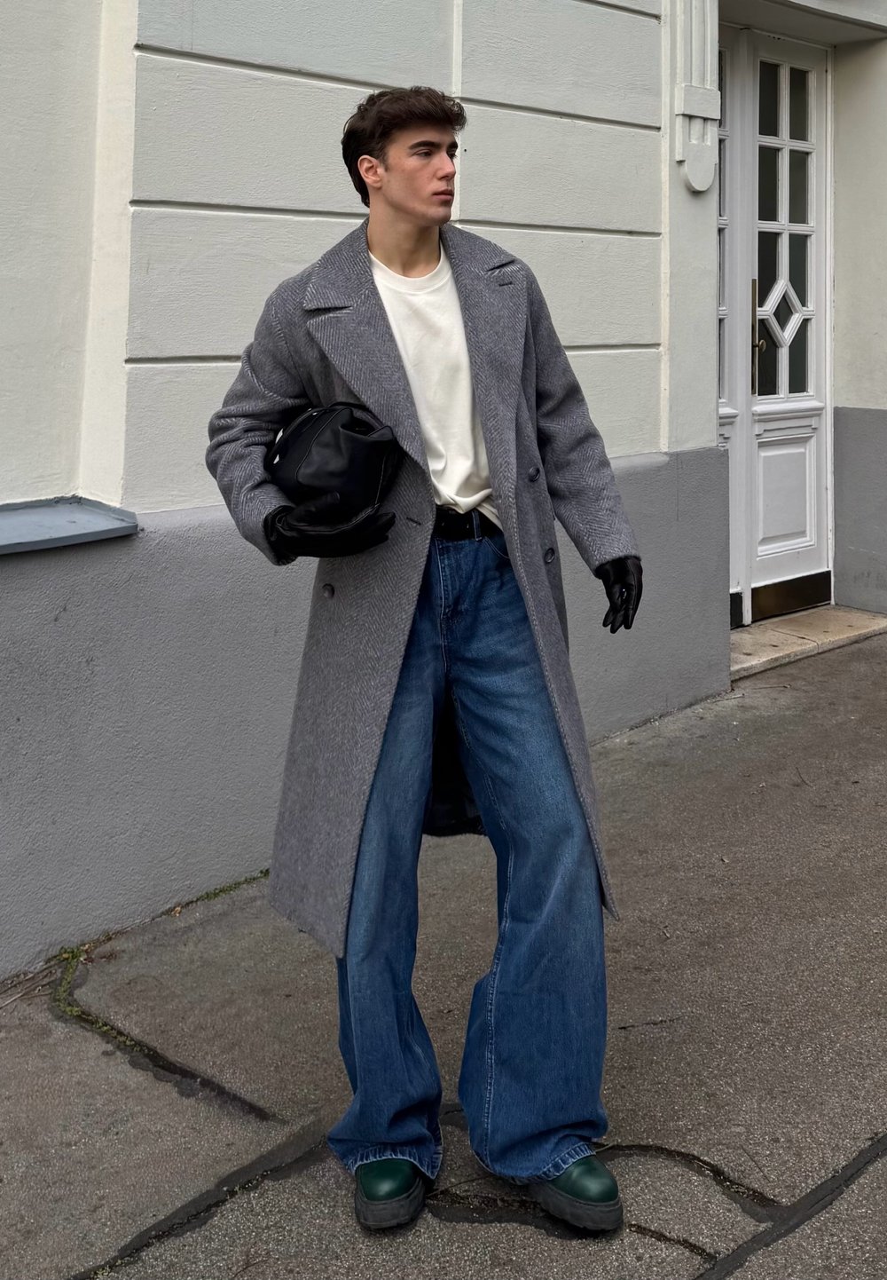 Sobretudo cinza com padrão espinha de peixe, t-shirt branca, jeans azul de perna larga, luvas pretas e sapatos verde escuro, segurando uma mala preta, em pé ao ar livre.