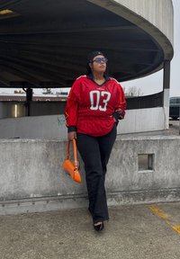 Červený dres s bielym číslom „03“, čierne rozšírené džínsy a oranžová kabelka. V pozadí je betónová konštrukcia a zamračená obloha.