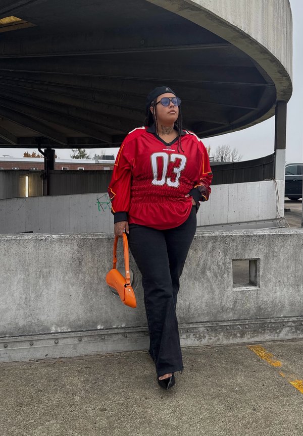 Rotes Trikot mit weißer "03", schwarze ausgestellte Jeans und eine orangefarbene Handtasche. Im Hintergrund ist eine Betonstruktur und ein bedeckter Himmel zu sehen.