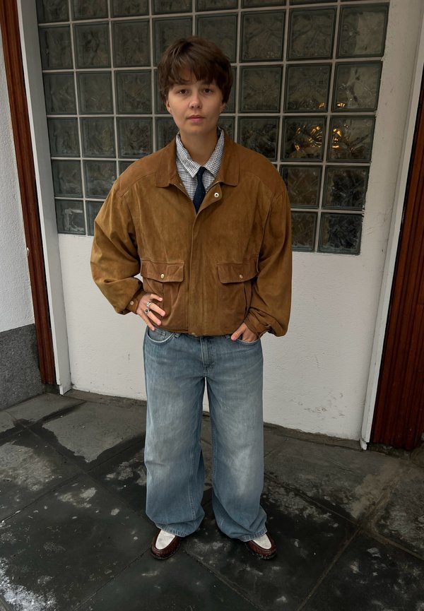 Personne aux cheveux bruns courts portant une veste en daim marron, une chemise à carreaux, une cravate foncée, un jean bleu ample et des chaussures marron et blanches, debout à l'intérieur.