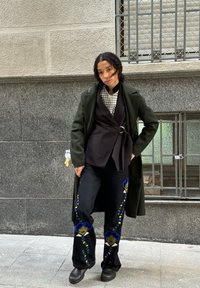 Zaļš vilnas mētelis pār melnu uzvalku, melni džinsi ar zilu ziedu izšuvumu un melnas zābaki. Gluda tekstūra ar slāņotu dizainu.