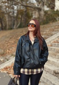 Sort læderjakke med løs pasform, lynlåslukning foran og skulderepauletter, lagdelt over en ternet skjorte og kombineret med store solbriller.