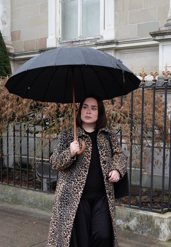Femme tenant un parapluie noir, portant un manteau à motif léopard, marchant sur un trottoir mouillé près d'une clôture en fer avec des plantes sèches.