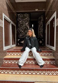 Giovane donna con occhiali e fascia, che indossa una giacca nera e pantaloni bianchi, seduta su gradini decorati con piastrelle in un ingresso con piastrelle marroni.