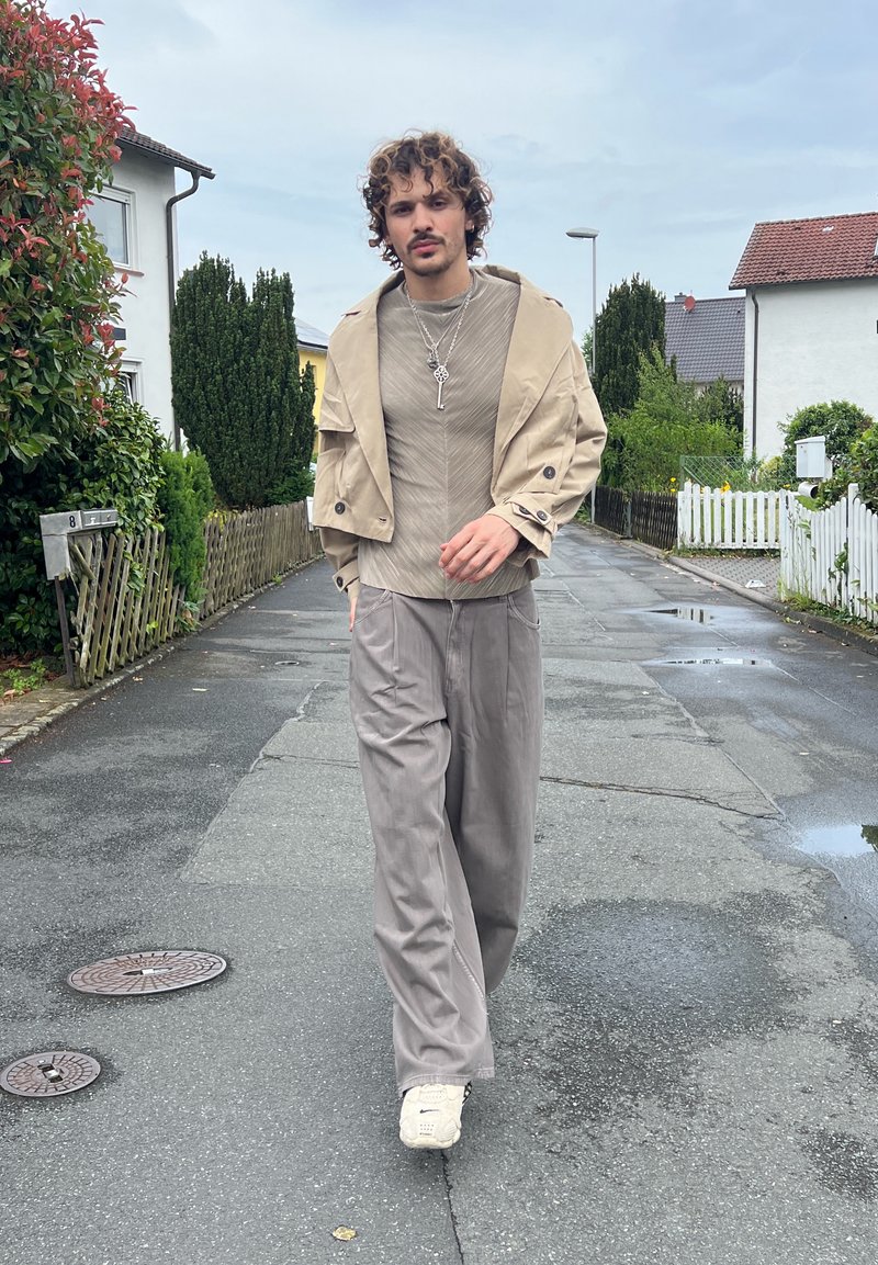 Homme aux cheveux bouclés marchant dans une rue mouillée, portant une veste beige, un pantalon gris, des baskets blanches et des colliers superposés.