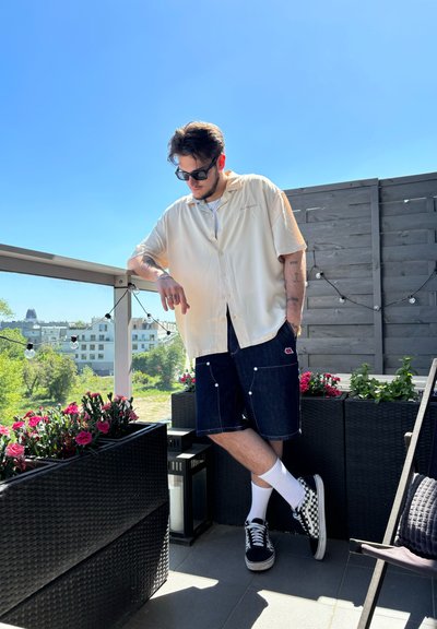 Camisa corta de manga en beige claro, pantalones cortos de mezclilla oscura con costuras blancas, zapatillas de cuadros en blanco y negro, de pie entre flores en un balcón.