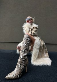 Mulher agachada, usando grandes óculos de sol marrons, um casaco bege forrado de pelagem e botas até ao joelho com padrão de pele de cobra, pousada contra uma parede cinza lisa.