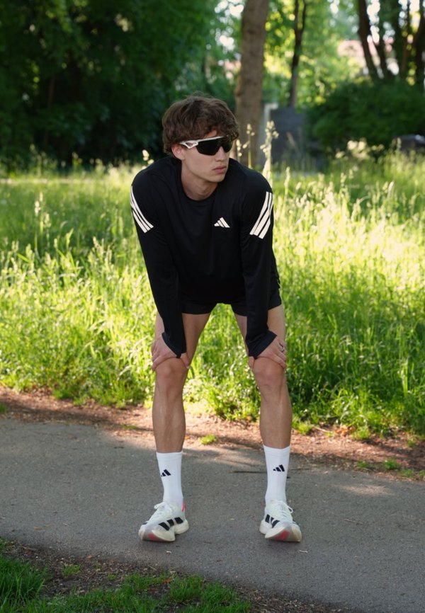 Jeune homme en tenue de sport noire et lunettes de soleil se tient sur un chemin du parc, penché en avant les mains sur les genoux, semblant se reposer après avoir couru.