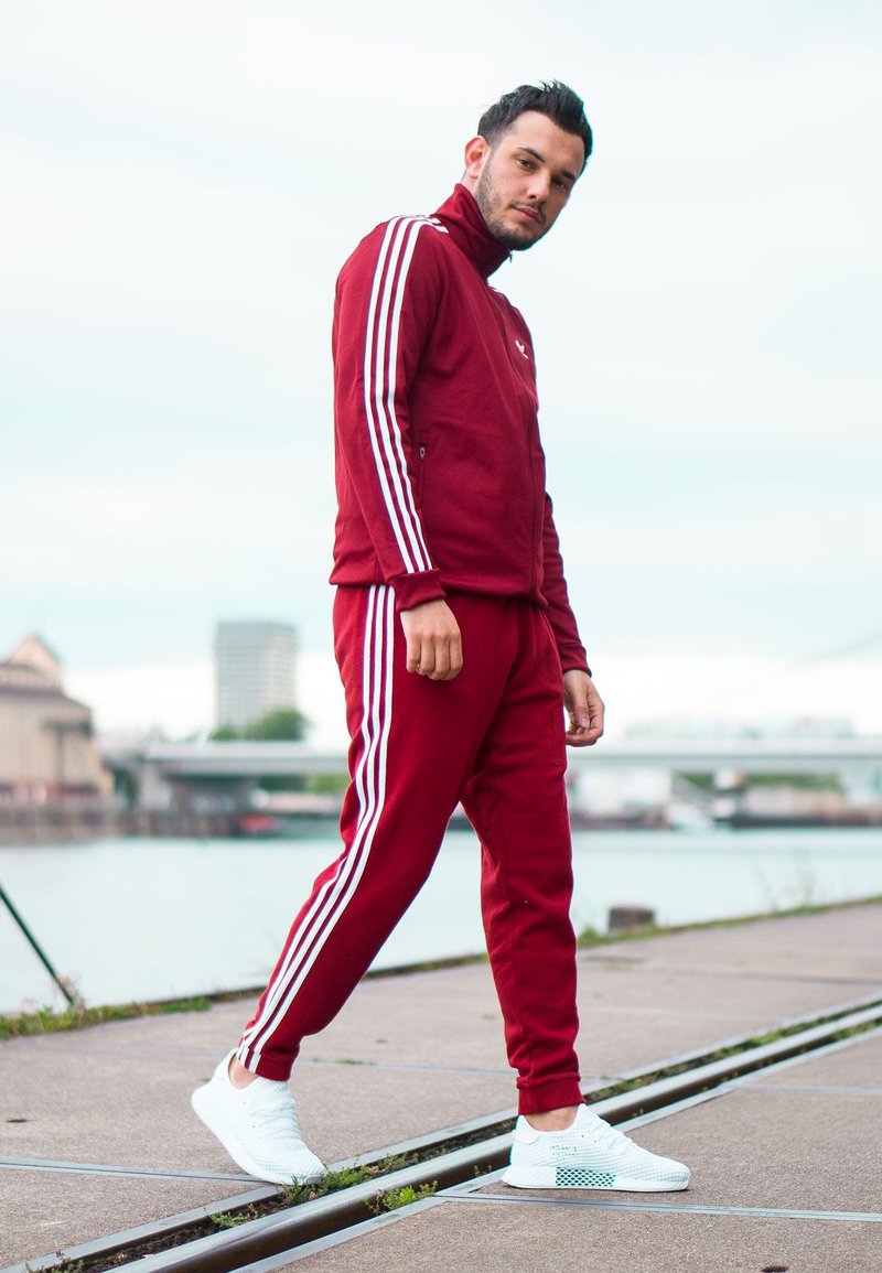 Roter Sportanzug mit weißen Streifen an den Ärmeln und Beinen, kombiniert mit weißen Turnschuhen. Der Stoff wirkt weich und hat ein figurnahes Design.