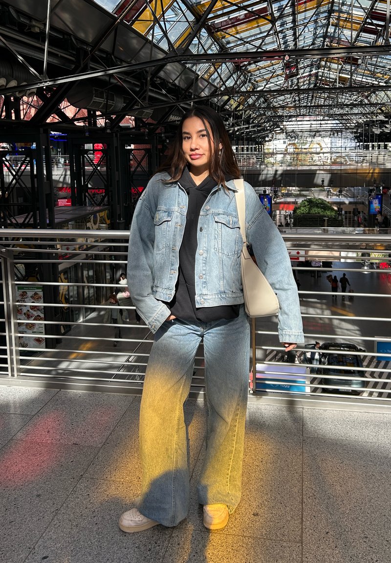 Uma jaqueta de denim azul claro sobre um moletom preto, combinada com calças de denim de perna larga em degradê. Ténis brancos e uma bolsa de ombro em creme.