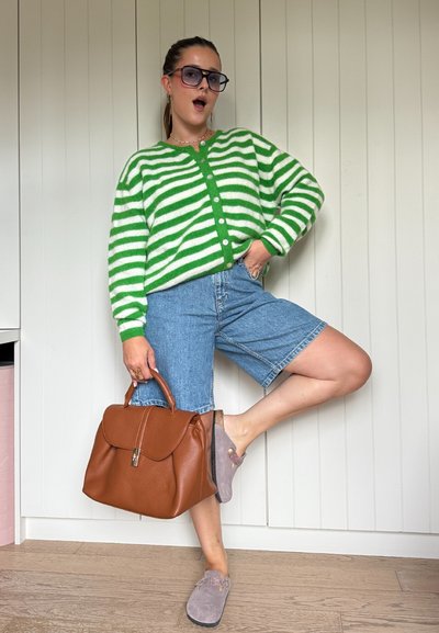 Cárdigan de punto a rayas verdes y blancas, pantalones cortos de mezclilla azules, bolso de cuero marrón, zapatos de gamuza gris con detalles, de pie contra un fondo blanco.