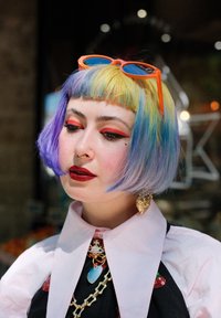 Une coupe de cheveux bob colorée et courte mettant en valeur des teintes pastel de jaune, violet et bleu. Les accessoires comprennent des lunettes de soleil oranges et des boucles d'oreilles en or.