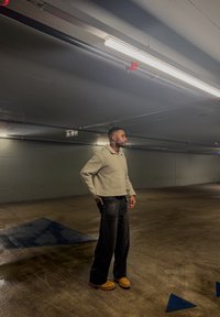 Homme portant un pull beige et un pantalon noir debout dans un parking vide faiblement éclairé avec des flèches directionnelles bleues au sol.