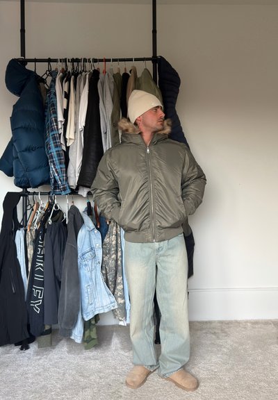 Hombre con gorro beige, chaqueta acolchada verde oliva, vaqueros azul claro y botas beige, de pie junto a un perchero con chaquetas y camisas.