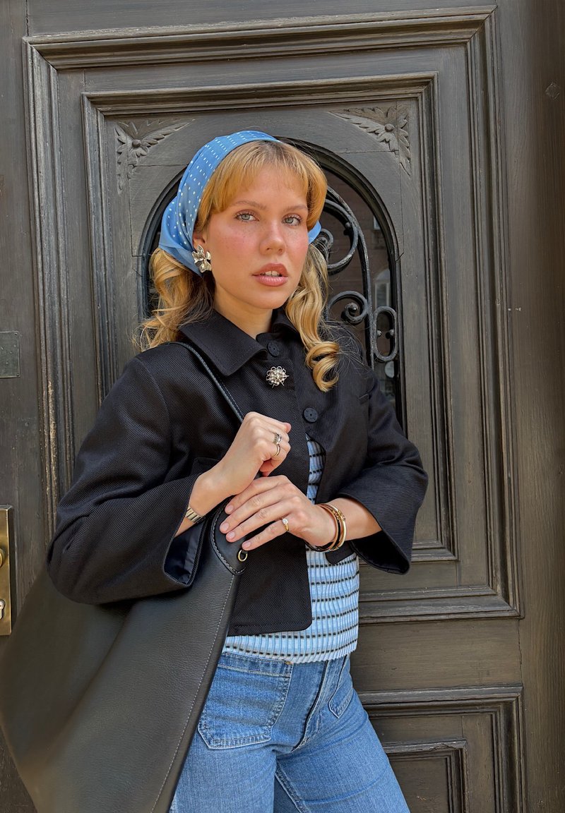 Jeune femme aux cheveux blonds et foulard bleu sur la tête, tenant un sac à main noir devant une porte en bois ornée.