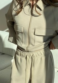 Beige zip-up top featuring two front pockets, paired with matching elastic-waist trousers. Smooth texture and relaxed fit. Natural light accentuates the fabric.