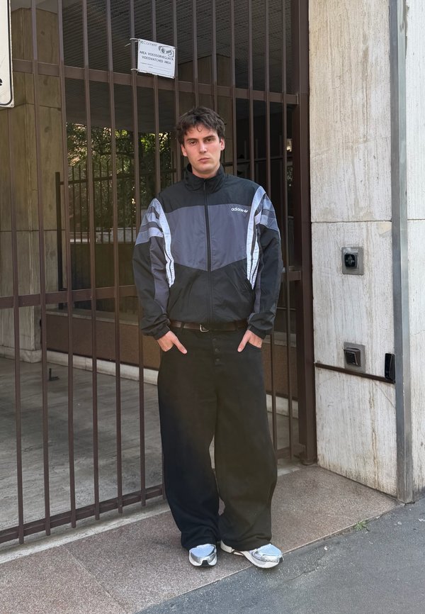 Giovane uomo in piedi con le mani nelle tasche, indossa una giacca Adidas nera e grigia, pantaloni neri larghi e sneakers argento vicino a un ingresso recintato.
