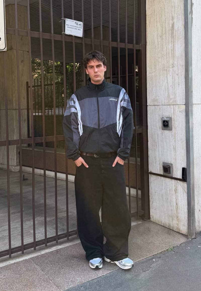 Jeune homme debout les mains dans les poches, portant une veste Adidas noire et grise, un pantalon noir ample et des baskets argentées près d'une entrée close.