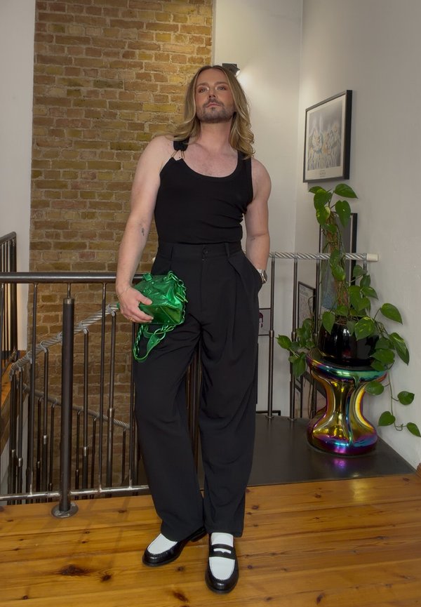 Persona con pelo largo, con camiseta sin mangas negra y pantalones, sostiene una bolsa verde, de pie en el interior cerca de una barandilla y un soporte para plantas colorido.