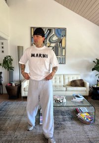 Λευκό μπλουζάκι με μαύρο κείμενο "MARNI", συνδυασμένο με γκρι φόρμες. Άτομο που φοράει μαύρη σκούφια, στέκεται σε φωτεινό δωμάτιο με έπιπλα.