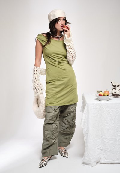 Mujer con vestido y pantalones verde oliva con mangas de crochet y boina, sosteniendo una manzana cerca de su boca, de pie junto a una mesa con frutas.