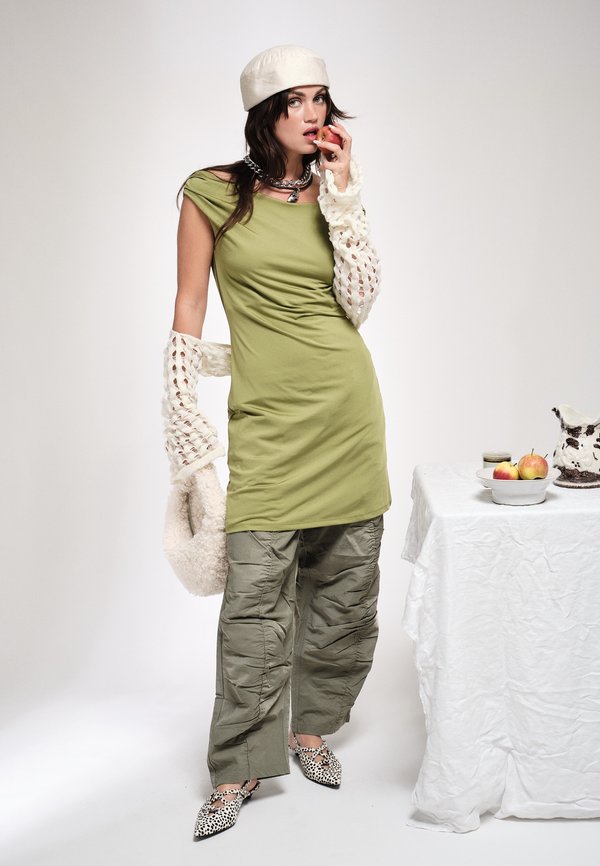 Γυναίκα με ελιάς πράσινο φόρεμα και παντελόνι με μανίκια κροσέ και μπερέ, κρατώντας ένα μήλο κοντά στο στόμα της, όρθια δίπλα σε ένα τραπέζι με φρούτα.