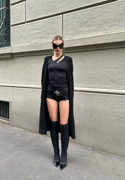 Cardigan largo negro sobre una camiseta de tirantes negra ajustada y pantalones cortos, con calcetines de canalé hasta la rodilla y tacones altos puntiagudos; unas grandes gafas de sol complementan el look.