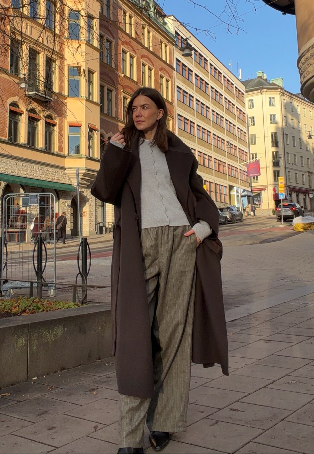 Longue veste marron portée sur un pull gris clair, assortie à un pantalon large à rayures fines. Boutons de la blouse et chaussures noires pointues visibles.