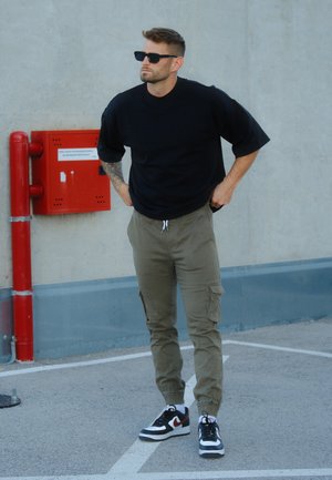 Camiseta negra oversize, pantalones cargo oliva con bolsillos laterales y zapatillas negras con detalles rojos. Atuendo urbano casual.