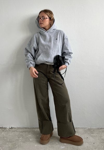 Sudadera de felpa gris con un bolsillo frontal, pantalones de pierna ancha en verde oliva y zapatillas de plataforma marrones; sosteniendo un pequeño bolso negro.