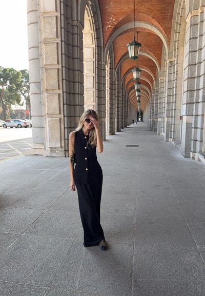 Vestido negro sin mangas con botones dorados, de forma fluida, combinado con un bolso negro, enmarcado en un arco de piedra con lámparas de luz con patrones.