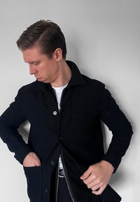 Veste noire avec un col, fermeture éclair à l'avant et bouton. Comprend une poche poitrine et des poches latérales. Texture lisse, coupe ajustée.