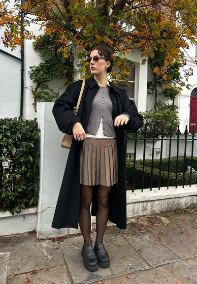 Abrigo largo negro, cárdigan gris, blusa crema, falda plisada de cuadros beige, medias negras y mocasines de plataforma. Accesorios incluyen gafas de sol y un bolso.