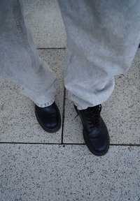 Zwarte leren laarzen met gele stiksels, gecombineerd met lichtgrijze, loszittende broeken, rustend op een gestructureerd stenen oppervlak.