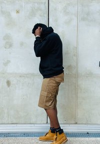 Mustandiga must kapuutsvi sweatshirt, pruunid cargo lühikesed püksid küljetaskutega ja kollased töösaapad, seistes tekstureeritud halli seina vastas.