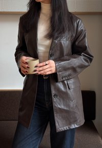 Personne portant une veste en cuir marron et un col roulé crème, tenant une tasse beige, debout à l'intérieur près d'un canapé marron.