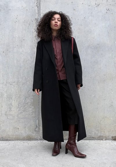 Joven con cabello rizado de pie contra una pared de cemento, vistiendo un abrigo largo negro, camisa de rayas, pantalones cortos y botas marrones con tacón.