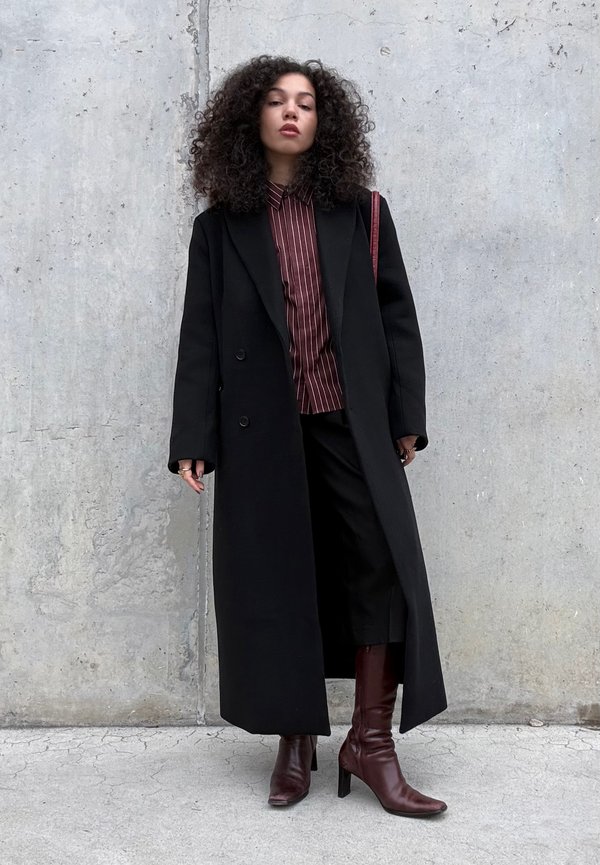 Junge Frau mit lockigem Haar, die vor einer Betonwand steht, trägt einen langen schwarzen Mantel, ein gestreiftes Hemd, gekürzte Hosen und braune Stiefel mit Absatz.