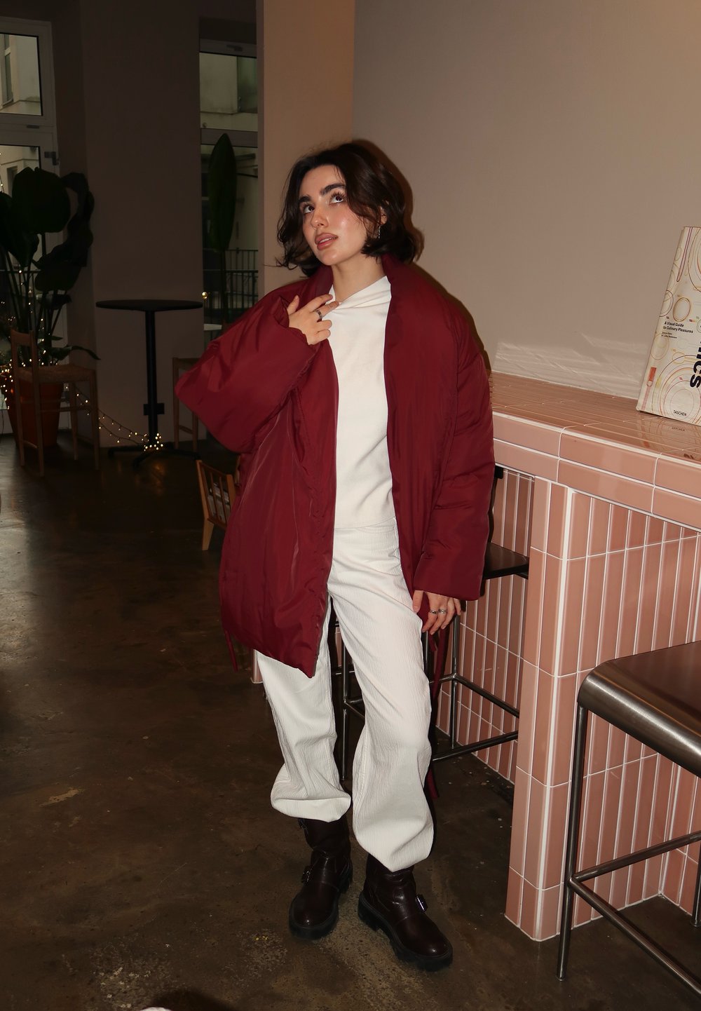 Burgundská prošívaná bunda přes bílý top, spárovaná s texturovanými bílými kalhotami a černými kotníkovými botami. Vnitřní prostředí s dekorativním pozadím.