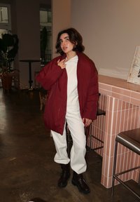 Burgundská prošívaná bunda přes bílý top, spárovaná s texturovanými bílými kalhotami a černými kotníkovými botami. Vnitřní prostředí s dekorativním pozadím.