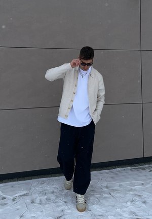 Hombre que lleva un cárdigan beige, camisa blanca, pantalones oscuros y gafas de sol, de pie sobre la nieve frente a una pared de azulejos marrones, ajustándose las gafas.