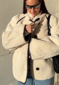 Femme portant une veste polaire crème avec des bordures noires, des lunettes de soleil foncées, des bagues argentées et un sac noir sur l'épaule.