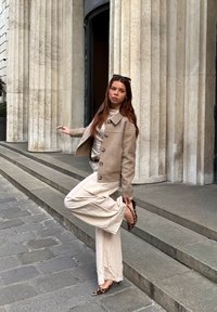Veste en laine beige avec des boutons sur le devant, blouse crème et pantalon large crème, associés à des escarpins à talon ouvert en imprimé léopard, devant des colonnes en pierre.