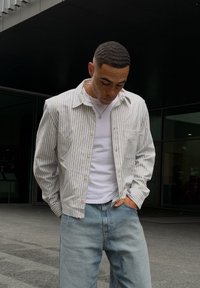 Camisa de botão às riscas em cores claras, combinada com uma t-shirt branca e jeans azul claro. Design casual, corte descontraído e mangas longas.