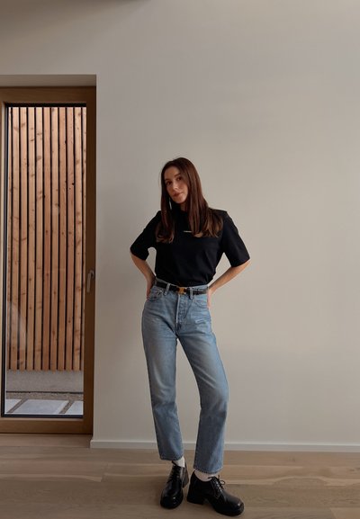 Top negro de manga corta, vaqueros de talle alto en azul claro, zapatos negros de cordones, de pie en un interior minimalista con acentos de madera.
