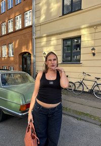Černý crop top s logem, vysokými tmavě modrými džíny a tkanou broskvovou kabelkou. V pozadí je vintage zelené auto a jízdní kola.