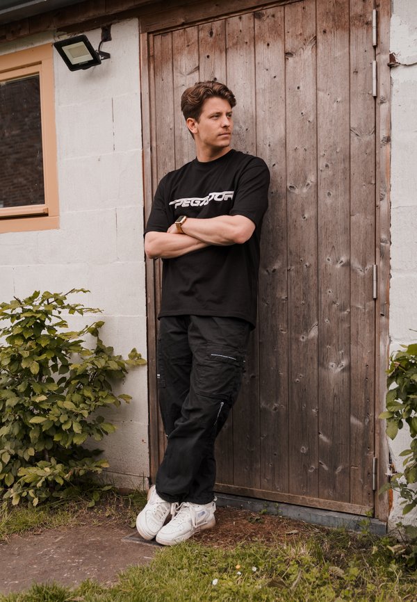 Camiseta negra con el logo "PEGATOR", pantalones cargo negros con bolsillos y zapatillas blancas. El fondo presenta una puerta de madera y vegetación.