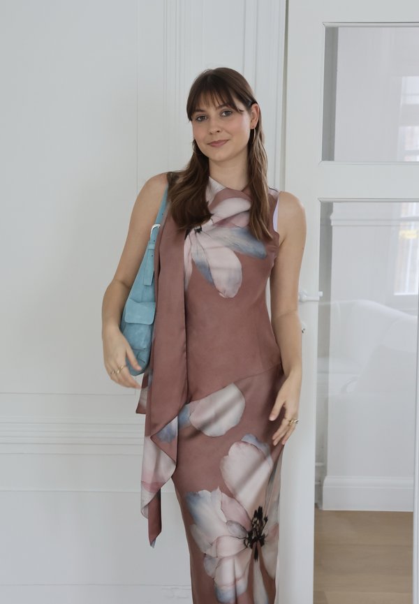 Jeune femme en robe sans manches mauve à motifs floraux, portant un sac à main bleu, debout à l'intérieur près d'un mur blanc et d'une porte vitrée.