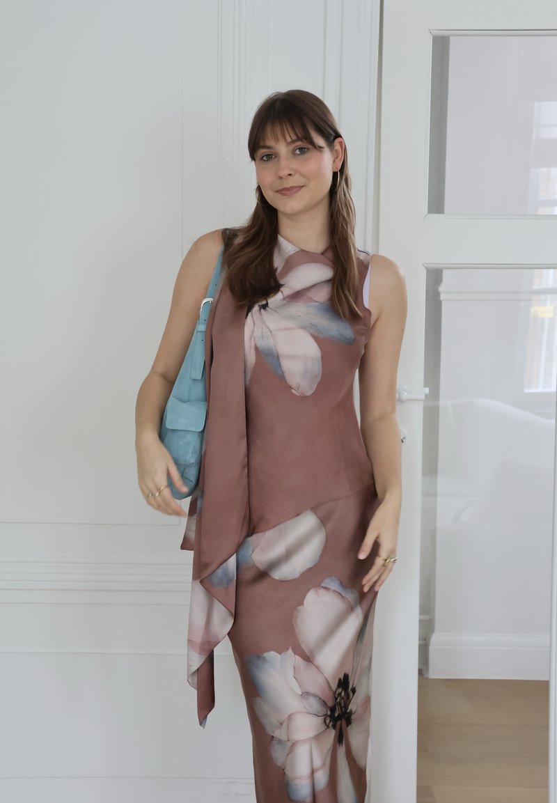 Jeune femme en robe sans manches mauve à motifs floraux, portant un sac à main bleu, debout à l'intérieur près d'un mur blanc et d'une porte vitrée.