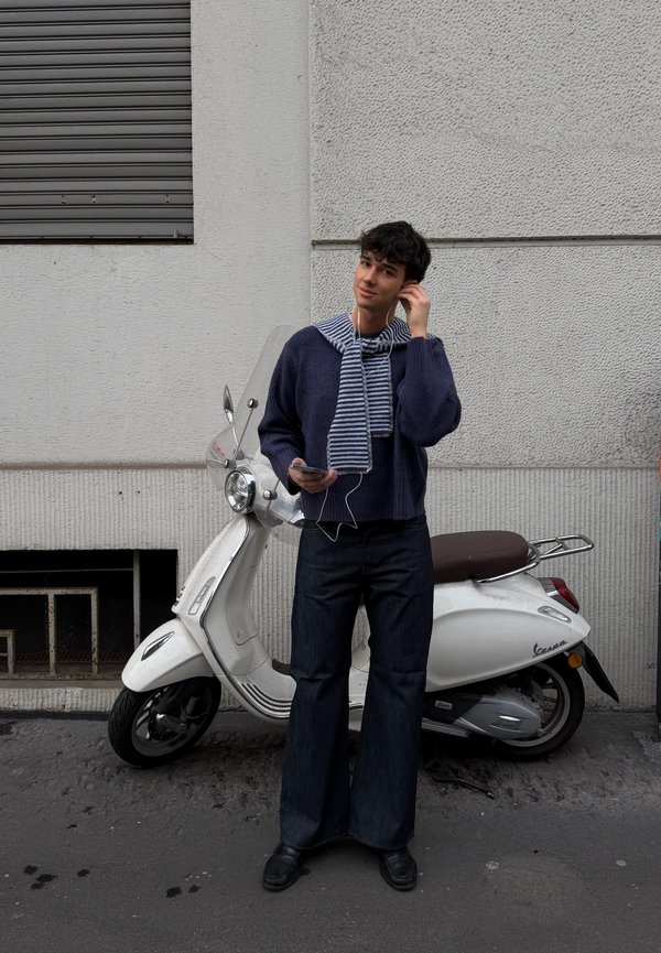Giovane uomo con maglione blu navy e sciarpa a righe, in piedi accanto a uno scooter bianco, mentre sistema gli auricolari tenendo un telefono in mano.